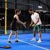 A coach shows a player how to hold the racket on a blue indoor padel court; she is wearing a black CRIVIT sports dress, he is wearing a white CRIVIT T-shirt and green CRIVIT shorts and is holding a yellow ball.