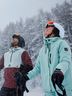 Two skiers in winter gear look up joyfully as snow falls in a forested mountain area.