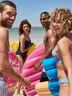 A group of four friends laughs and smiles on a beach, holding colorful inflatable floats.