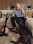 Andre Agassi siedzi na kanapie i reaguje żartobliwie, podczas gdy duży pies siedzi pośród porozrywanego wypełnienia na dywanie; obok stoi bezprzewodowy odkurzacz SILVERCREST w przytulnym salonie.