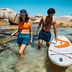 Un hombre y una mujer se encuentran en aguas poco profundas junto a una tabla de paddle surf, disfrutando de un día soleado junto a las rocas.