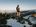 Woman in CRIVIT outdoor outfit with rucksack stands on snow-covered mountain peak with sweeping panoramic view.