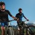 Dwóch mężczyzn w stroju rowerowym CRIVIT jadących na rowerach górskich po alpejskiej drodze
