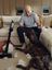 Andre Agassi sitting on a sofa, reacting playfully as a large dog sits amid torn stuffing on the carpet, with a cordless SILVERCREST vacuum cleaner nearby in a cozy living room.