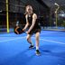Padel player in black CRIVIT sports dress stands in hitting position, ready for a backhand, on a blue indoor court, holding a black and orange CRIVIT racket; glass walls and fellow players in the background.