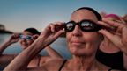 Woman adjusting black CRIVIT swim goggles on a rocky shoreline