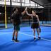 Two padel players high-five each other on a blue indoor court; the female player is wearing a black CRIVIT sports dress and holding a CRIVIT padel racket, while the male player is wearing a black CRIVIT shirt and green CRIVIT shorts.