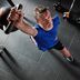A man works out in a gym, reaching forward with a weight or training handle. The overhead angle highlights the movement and intensity of the exercise. Gym flooring and equipment are visible around him.
