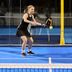 Padel player in black CRIVIT sports dress hits a forehand shot on a blue indoor court; yellow ball flies in front of black and orange CRIVIT racket, glass wall, and net in the background.