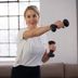 Frau in weißem CRIVIT-Sportshirt macht ein Schultertraining mit zwei CRIVIT-Kurzhanteln und streckt einen Arm seitlich aus; heller Raum mit Fenster im Hintergrund.