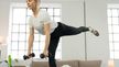 Fitness expert Linda performs a single‑leg bend with two CRIVIT dumbbells in a bright living room.