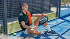 Young man resting on a padel court fence holding a CRIVIT padel racket and wearing a black polo shirt with orange towel over his shoulder.