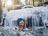 Woman with hat smiles as she bathes in an ice-cold waterfall in a winter forest.
