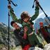 A group hikes through rocky mountain terrain under a clear blue sky. A child in front holds trekking poles while others follow with backpacks. The scene suggests an outdoor adventure trip.