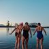 Drie vrouwen in badkleding lopen samen een rustig meer in, omgeven door natuur.