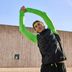 L'expert Tim fait un étirement du torse debout avec les bras tendus vers le haut devant une façade en bois, en tenue de course CRIVIT noire et vert fluo.