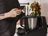 Person standing in a kitchen using a smartphone to control the black Monsieur Cuisine compact multifunctional cooking appliance via Monsieur Cuisine app.