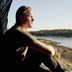 Erik Sander sits on a rock by the water, gazing into the distance – a forested shoreline and blue sky in the background.