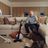 Andre Agassi sitting on a sofa, reacting playfully as a large dog sits amid torn stuffing on the carpet, with a cordless SILVERCREST vacuum cleaner nearby in a cozy living room.