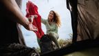 Woman in CRIVIT outdoor jacket handing gear out of a tent at a campsite